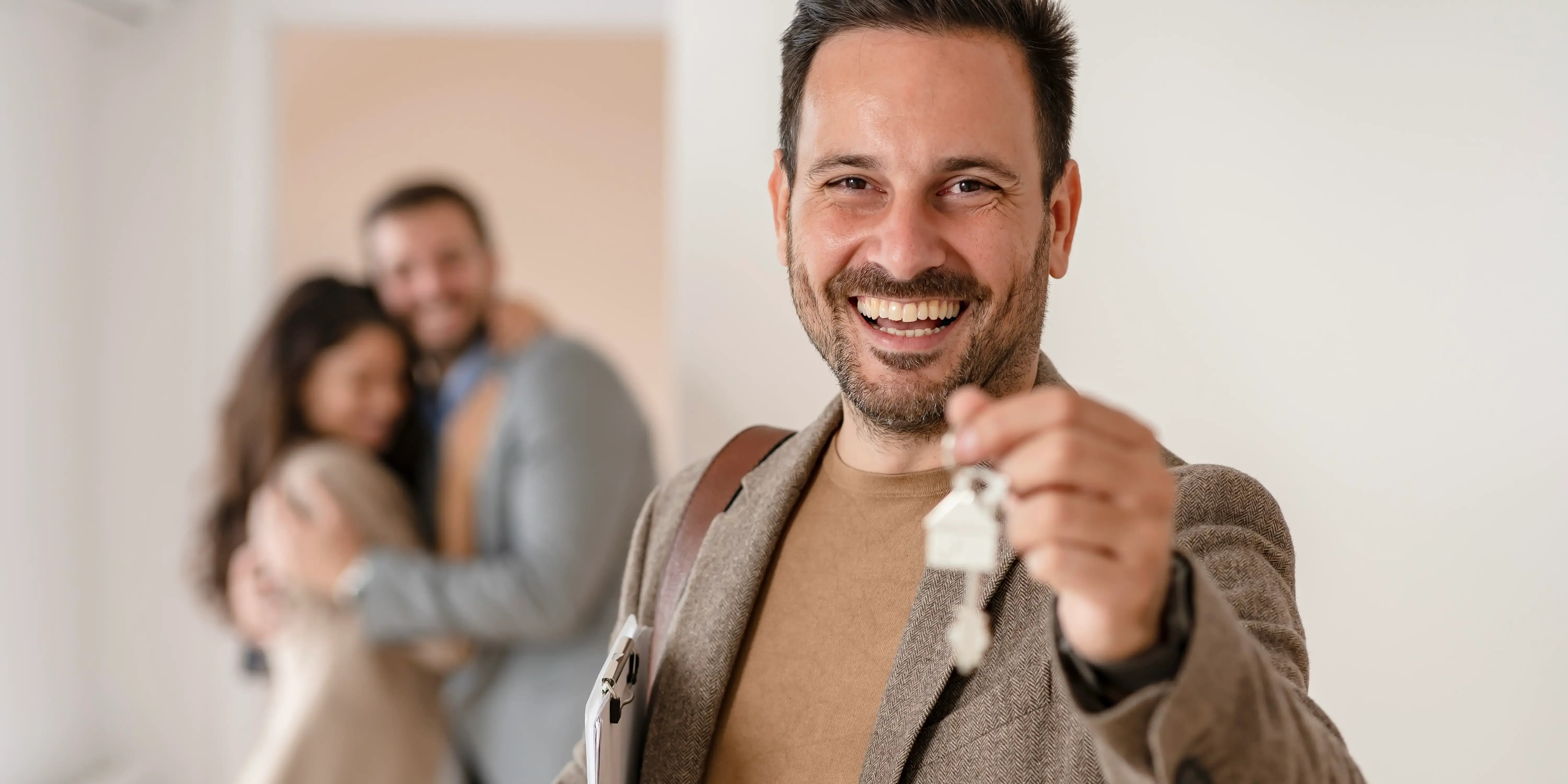 Asesor inmobiliario sosteniendo llaves de una casa económica, al fondo se ve borrosa una pareja abrazada y feliz de que acaba de comprar una casa