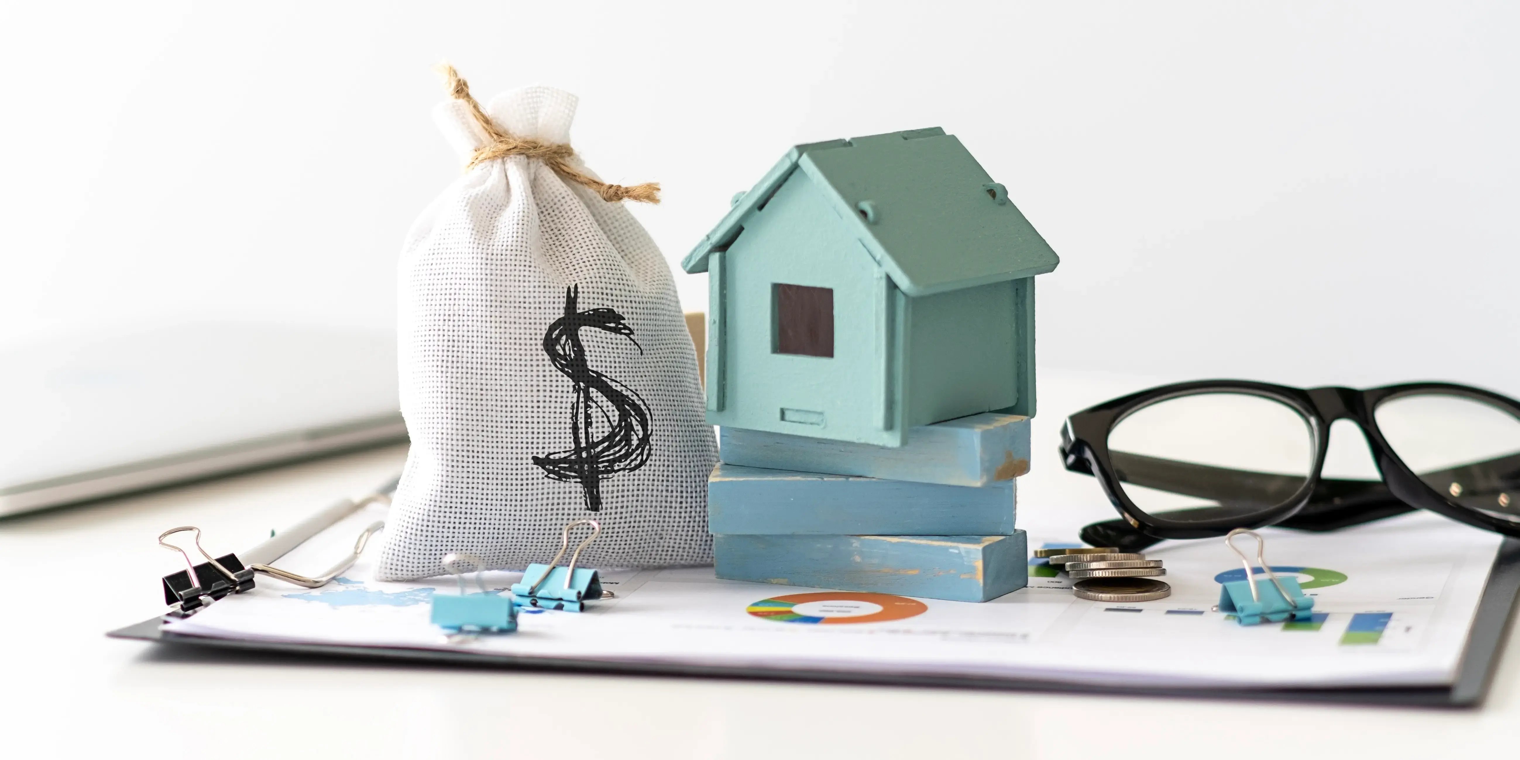 Casa de madera con una bolsa con signo de pesos y al otro lado unos lentes, todo sobre unas gráficas de papel de que se tiene una buena planeación financiera para comprar casa