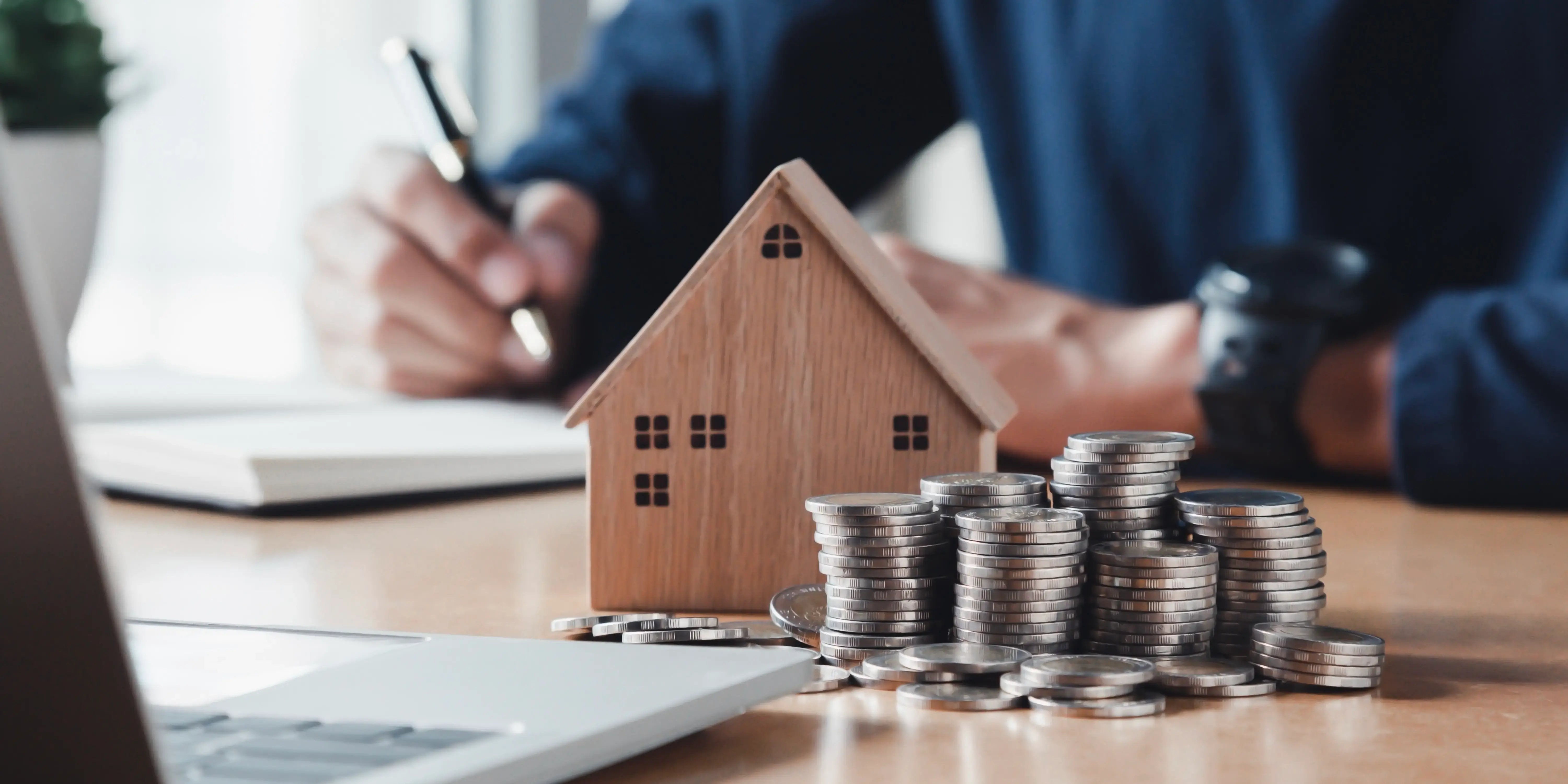 Casa de madera sobre una mesa con torres de monedas al frente representando la plusvalía que ha adquirido a través del tiempo