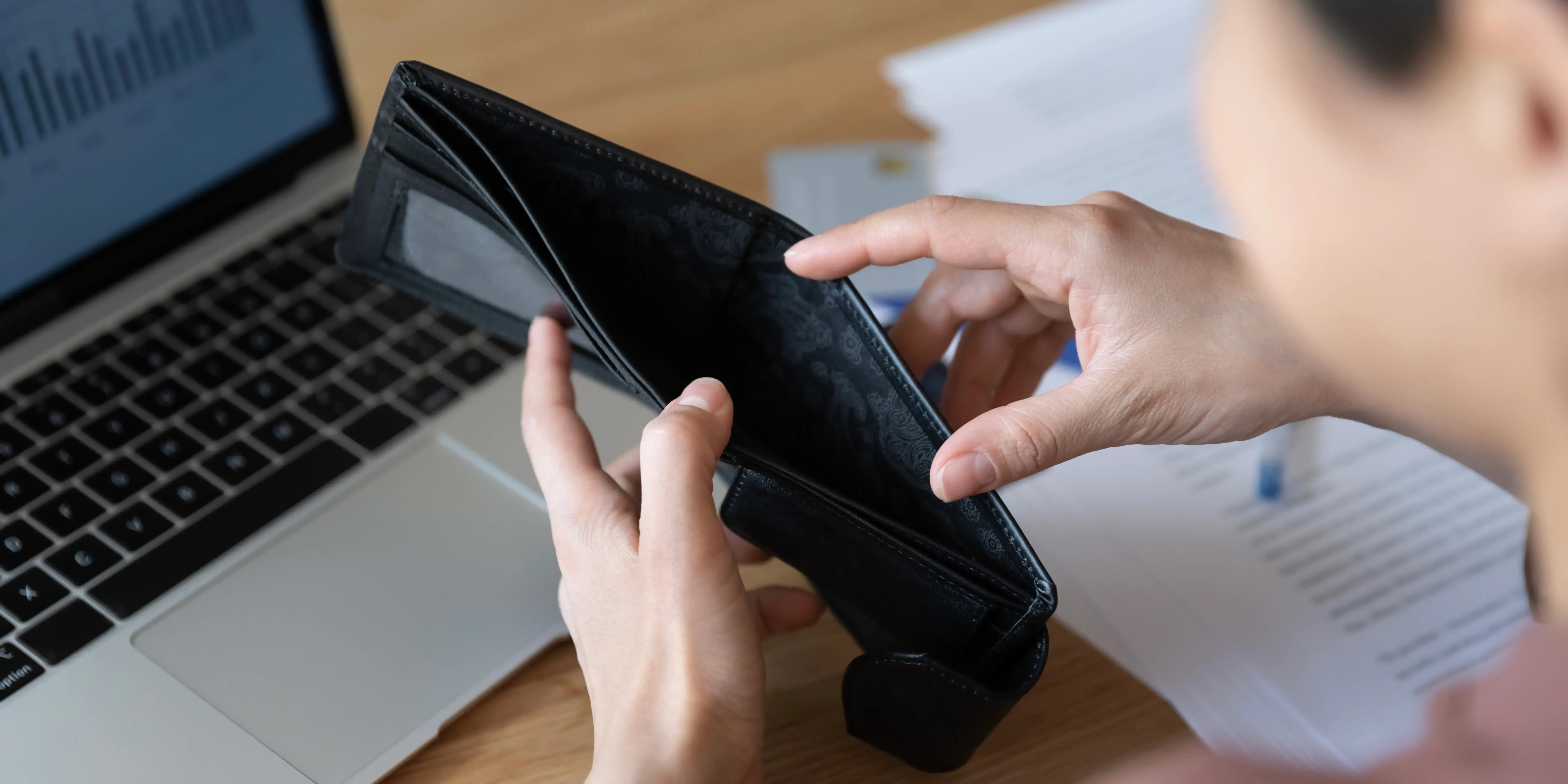 Hombre sosteniendo una cartera sin dinero, al fondo se ve una computadora con gráficas del dinero ahorrado