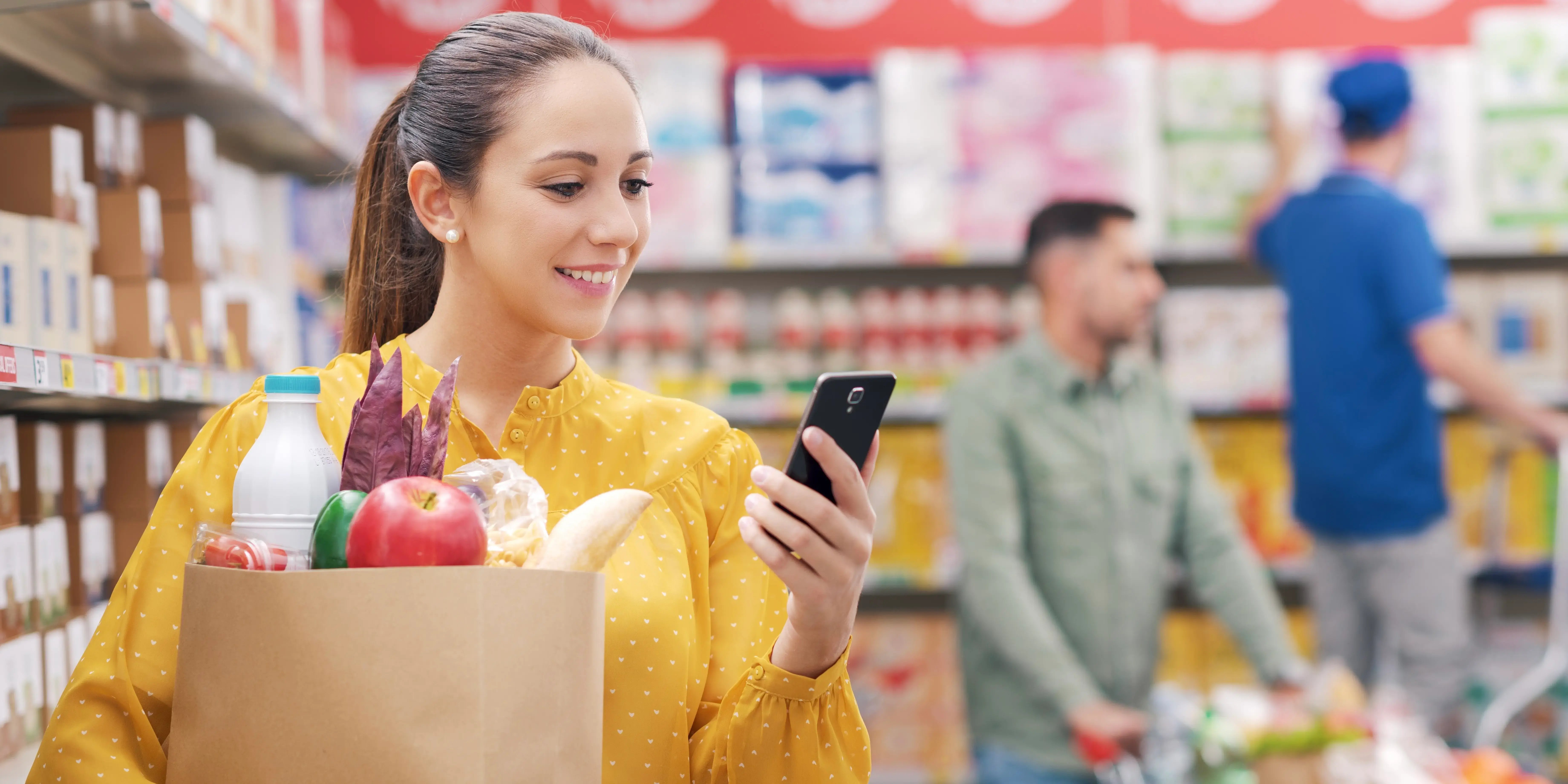 Mujer con poder adquisitivo haciendo compras en el supermercado, está sosteniendo una bolsa y haciendo cuentas con la calculadora del smartphone