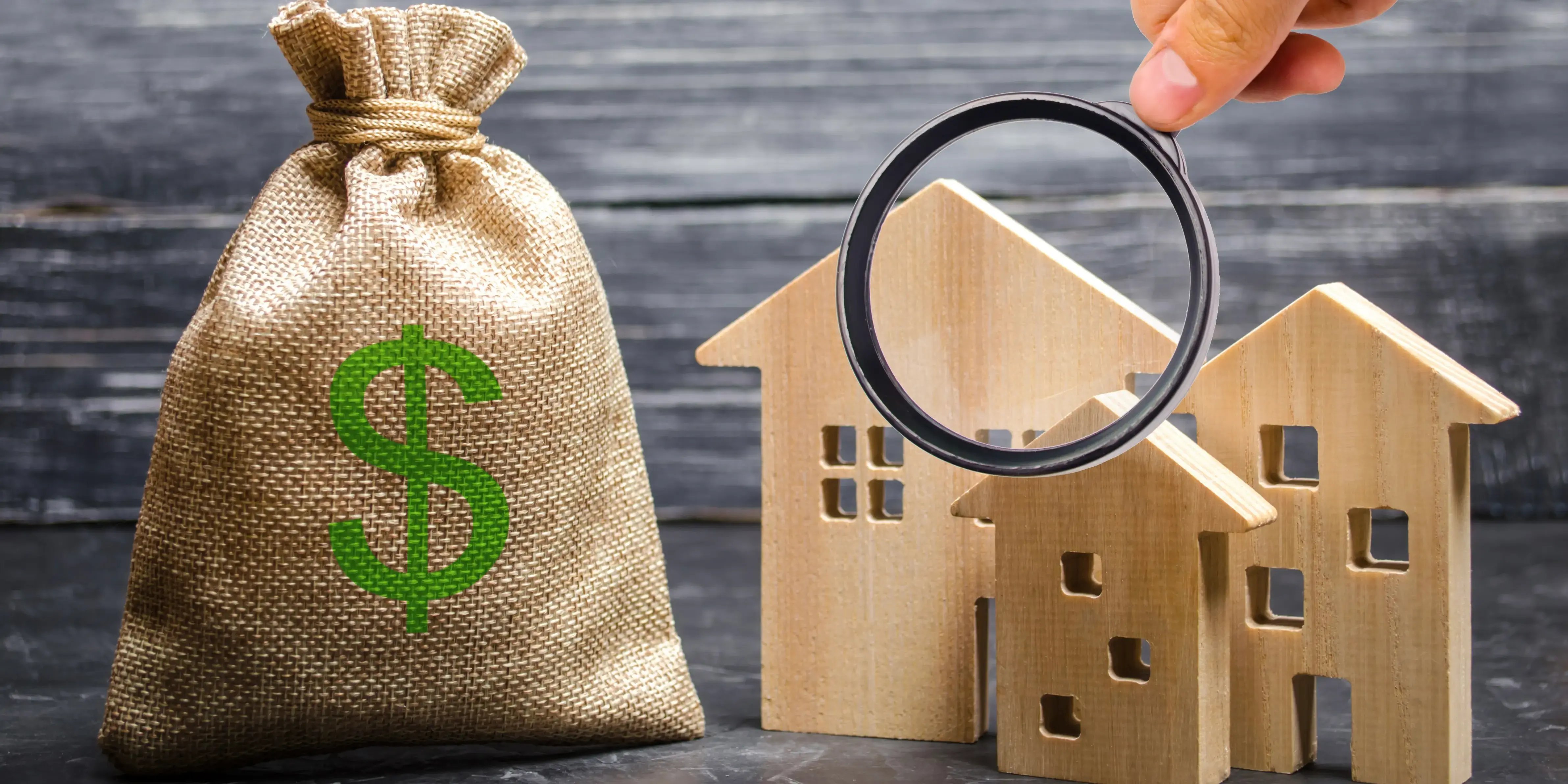 Persona sosteniendo una lupa y está mirando tres casas de madera cerca de una bolsa con dinero para concer el valor de las propiedades para elegir la mejor ubicación para vivir de acuerdo a su plusvalía