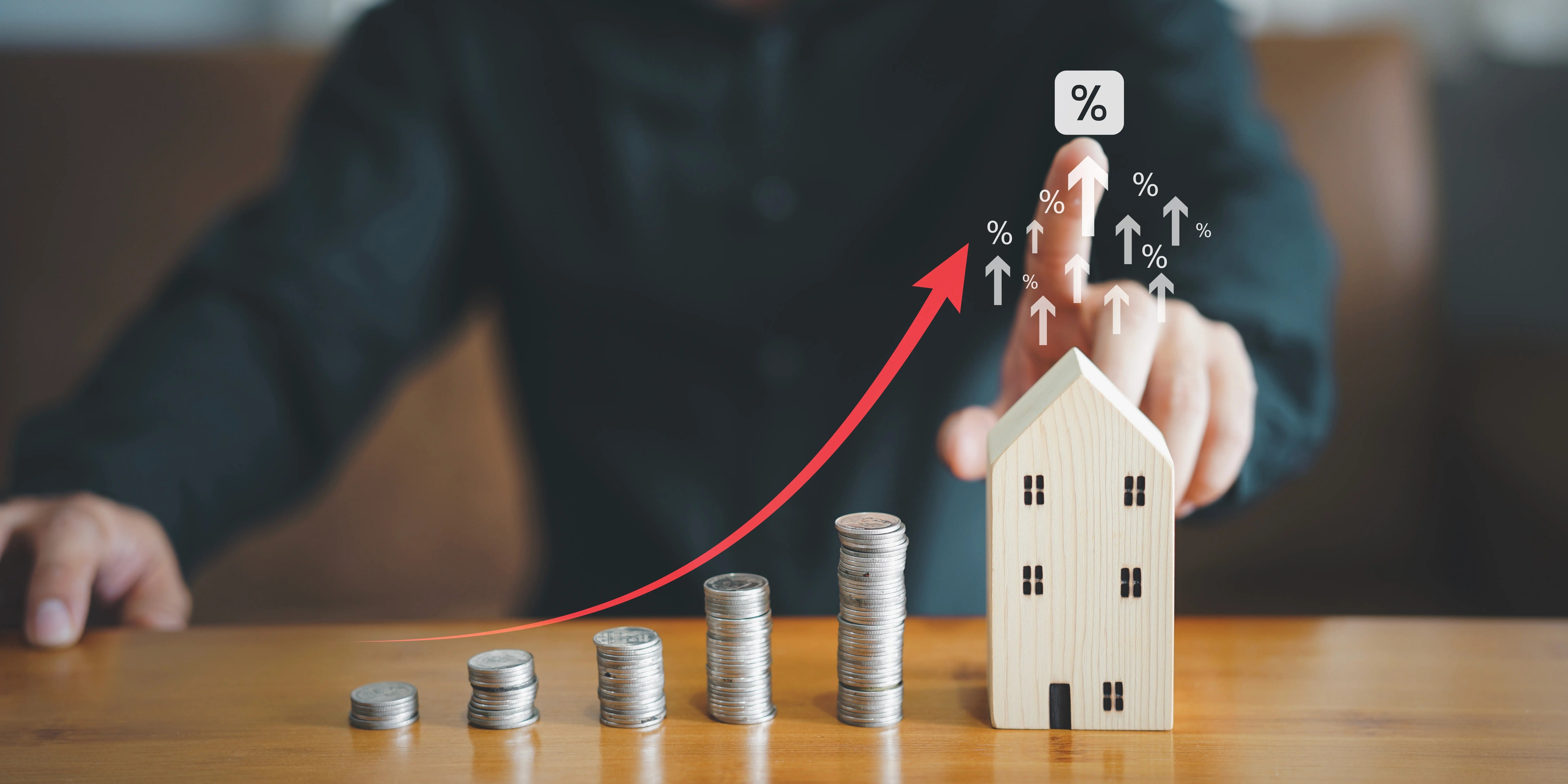 Torres de monedas en aumento y a lado una casa de madera donde un hombre está señalando un signo de porcentaje de que se pagarán mpas intereses al comprar una casa sin dinero ahorrado