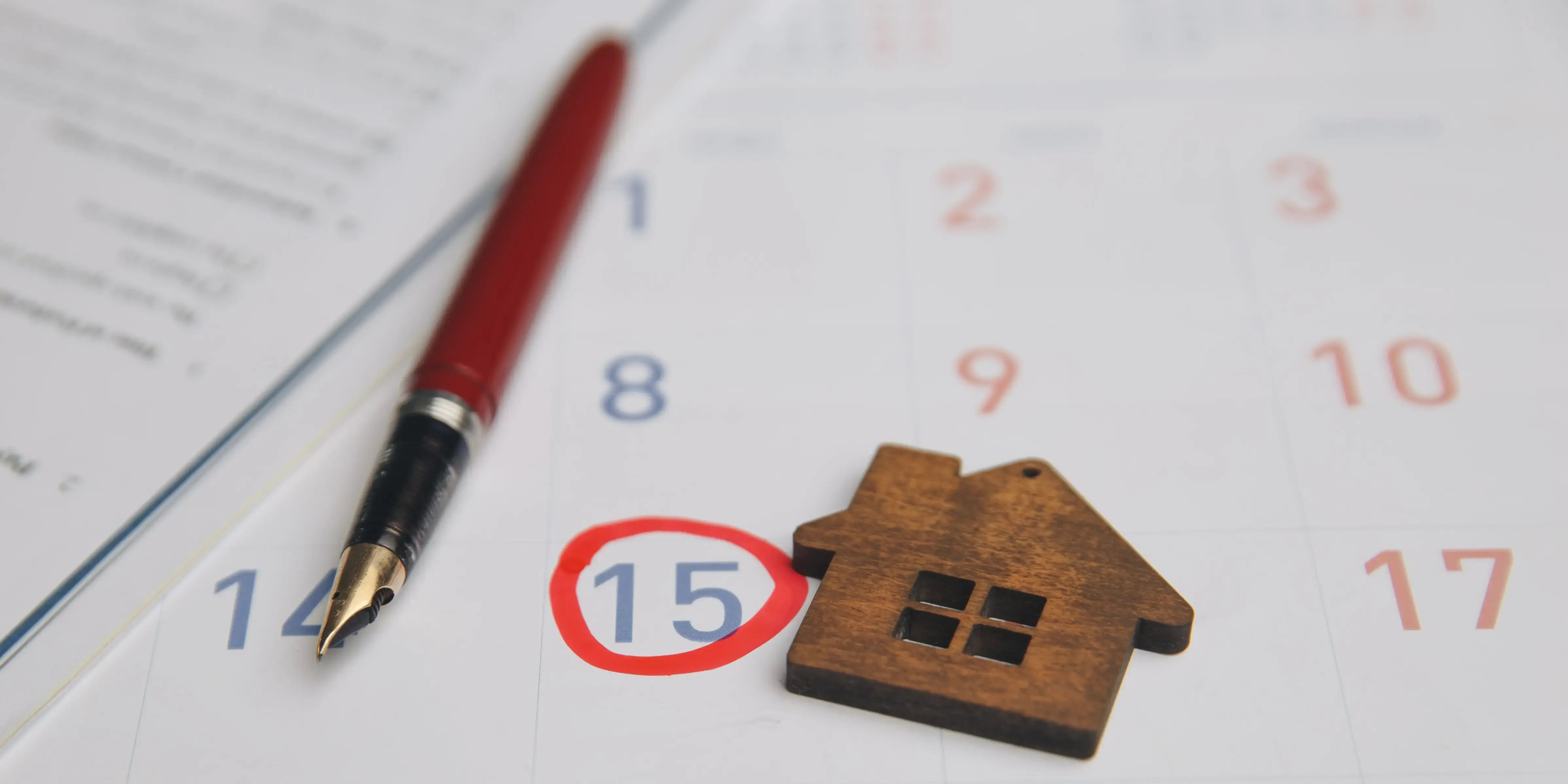 Una pluma y una casa de madera sobre un calendario señalando el día 15, día de pago del préstamo hipotecario para no generar intereses moratorios