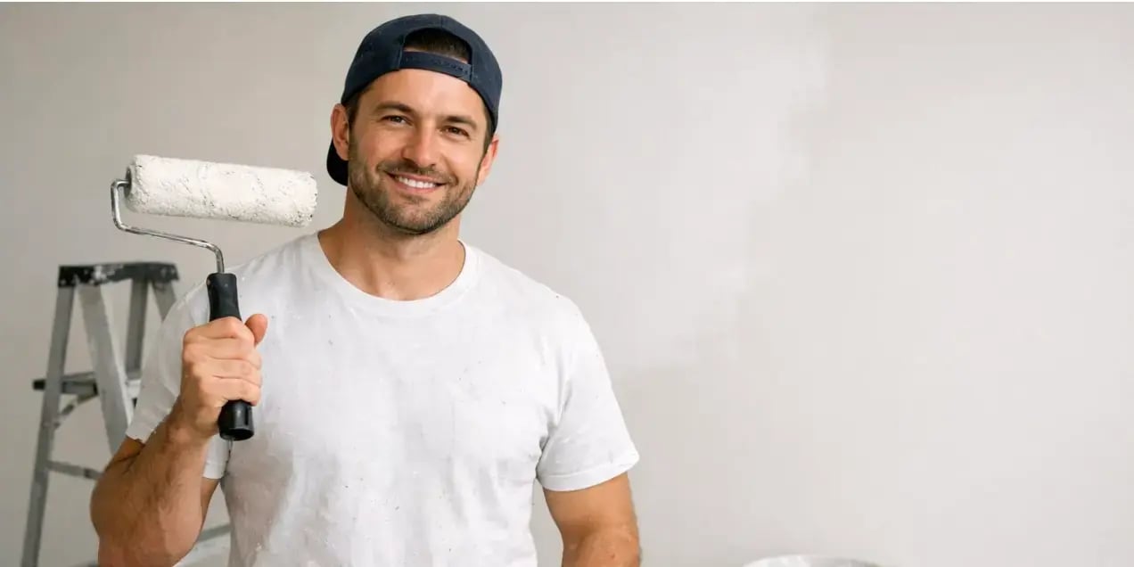 hombre en una sala sin muebles solo rodillo escalera y una cubeta de pintura