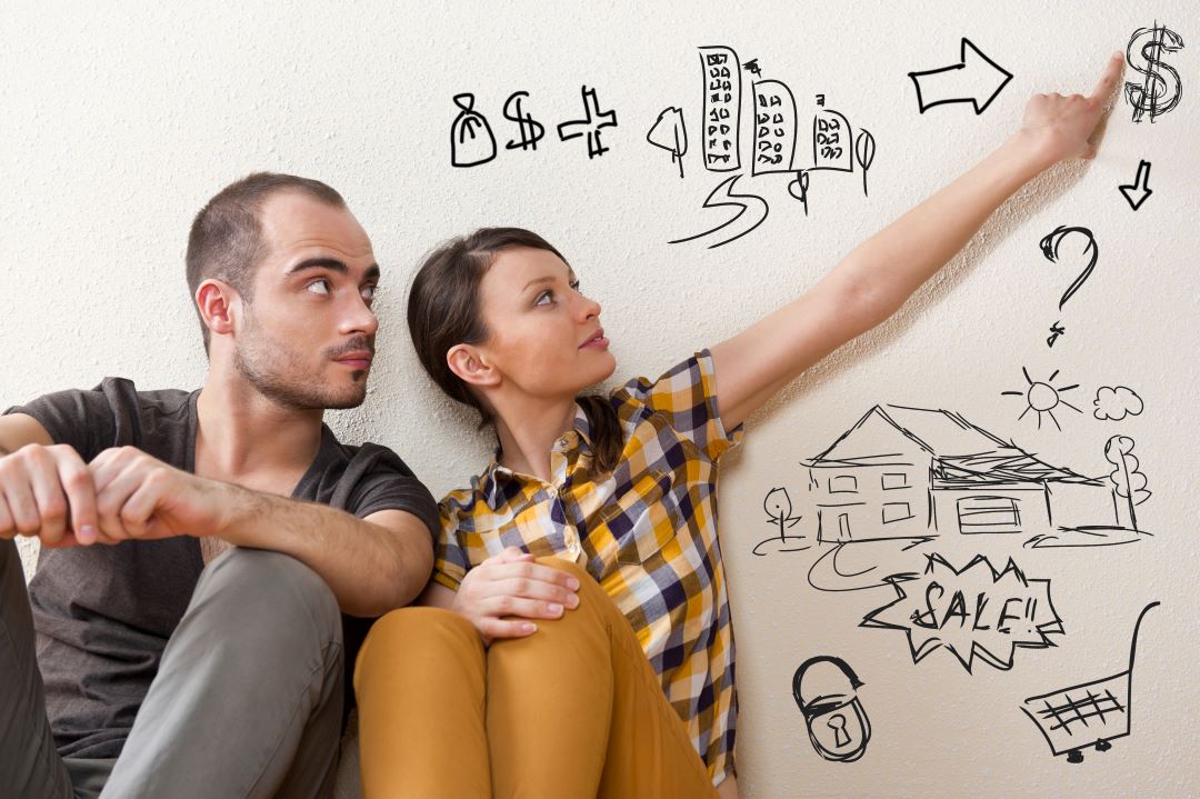 Una pareja de adultos jóvenes, están dibujando planes para comprar una casa en una pared blanca. Están sentados uno junto al otro. 