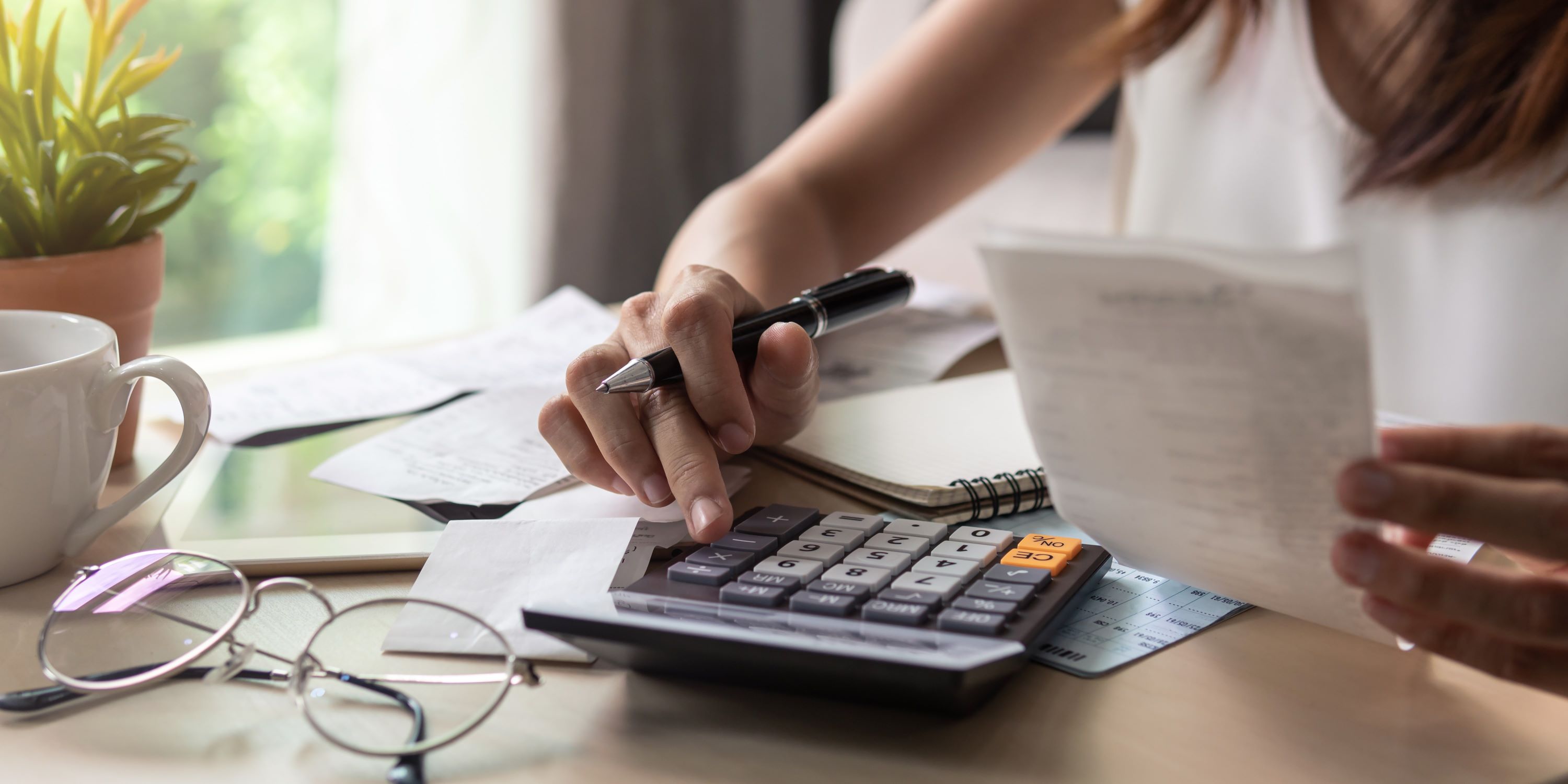 Mujer calcula su capacidad máxima de pago, tiene una calculadora, lentes y facturas en su escritorio.