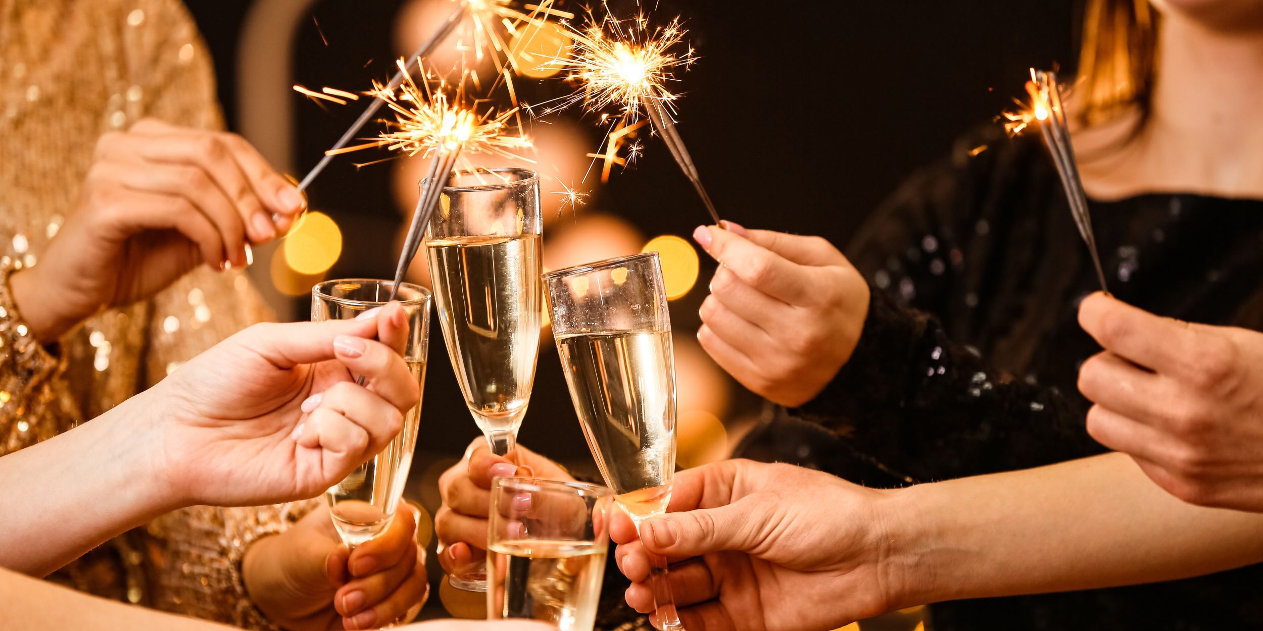 Año nuevo. Cuatro personas celebran con una copa y barita de chispas. Sus manos y antebrazos están al centro de la imagen. 