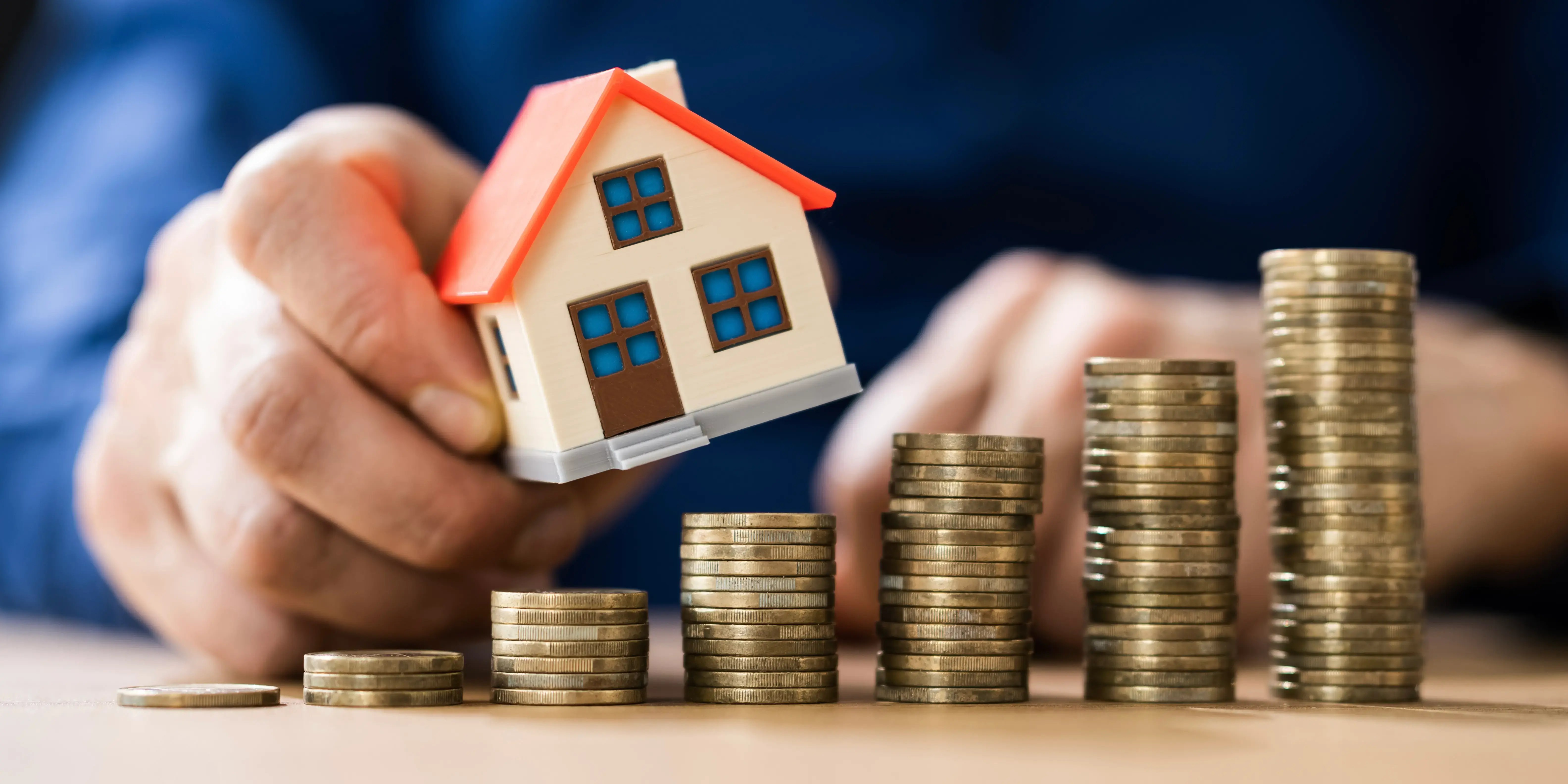 Hombre sosteniendo una casa de madera sobre torres de monedas para invertir en bienes raíces con poco dinero.