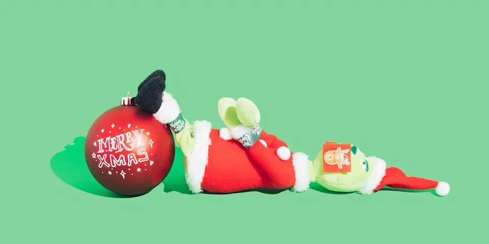 Un peluche con la figura del Grinch está maniatado, sus piecitos están en cima de una esfera navideña que dice Merryxmas. El fondo es verde.