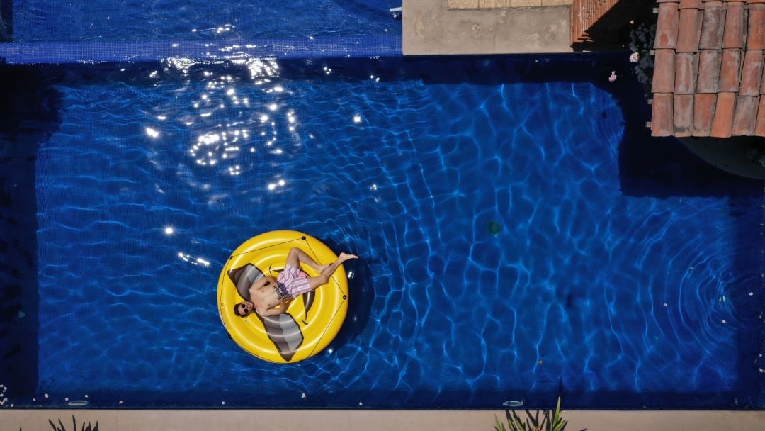 Hombre tomando el sol encima de un flotador con figura de emoji. En la esquina de la imagen hay un techo de tejas.